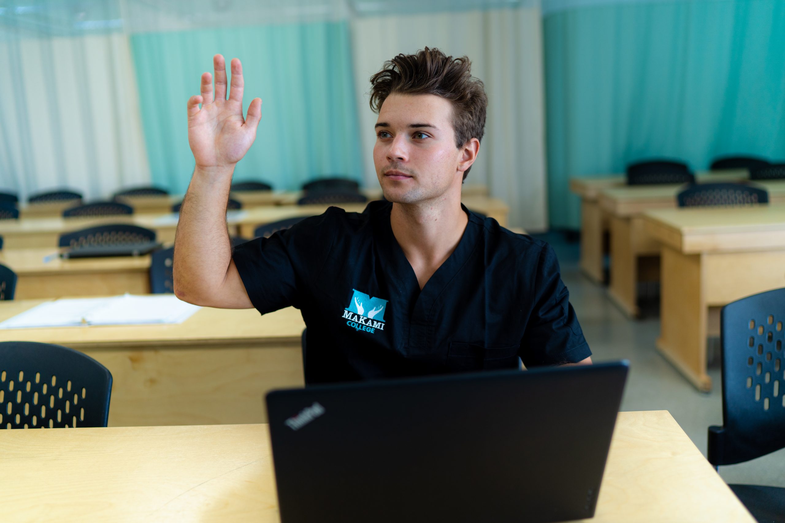 A young male MaKami College student raises his hand in an classroom while behind a black lap top