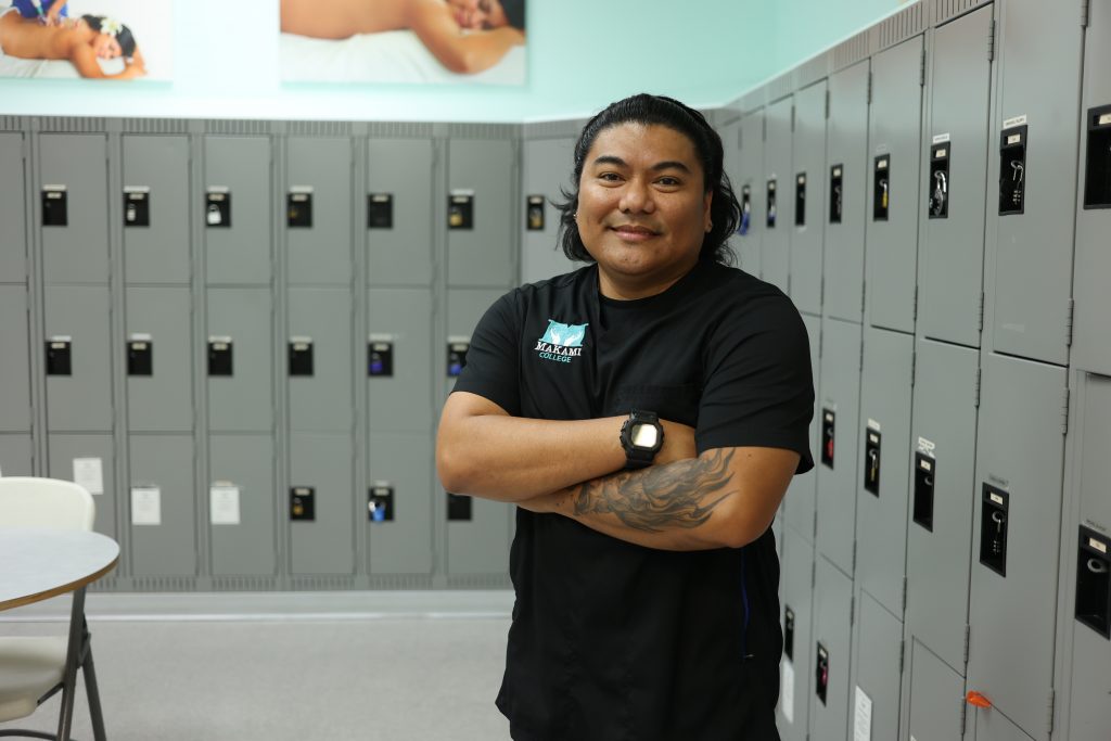 Derrol, a Health Care Aide graduate from MaKami College, stands in front of school lockers in a black scrub with his arms crossed.