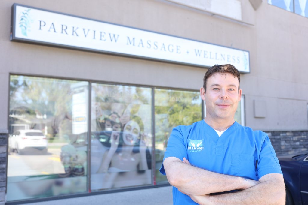 Donald, a caucasian male in his 30's, stands in front of his workplace Parkview Massage + Wellness in Calgary, Alberta, where he works as a massage therpaist while going to school at MaKami College