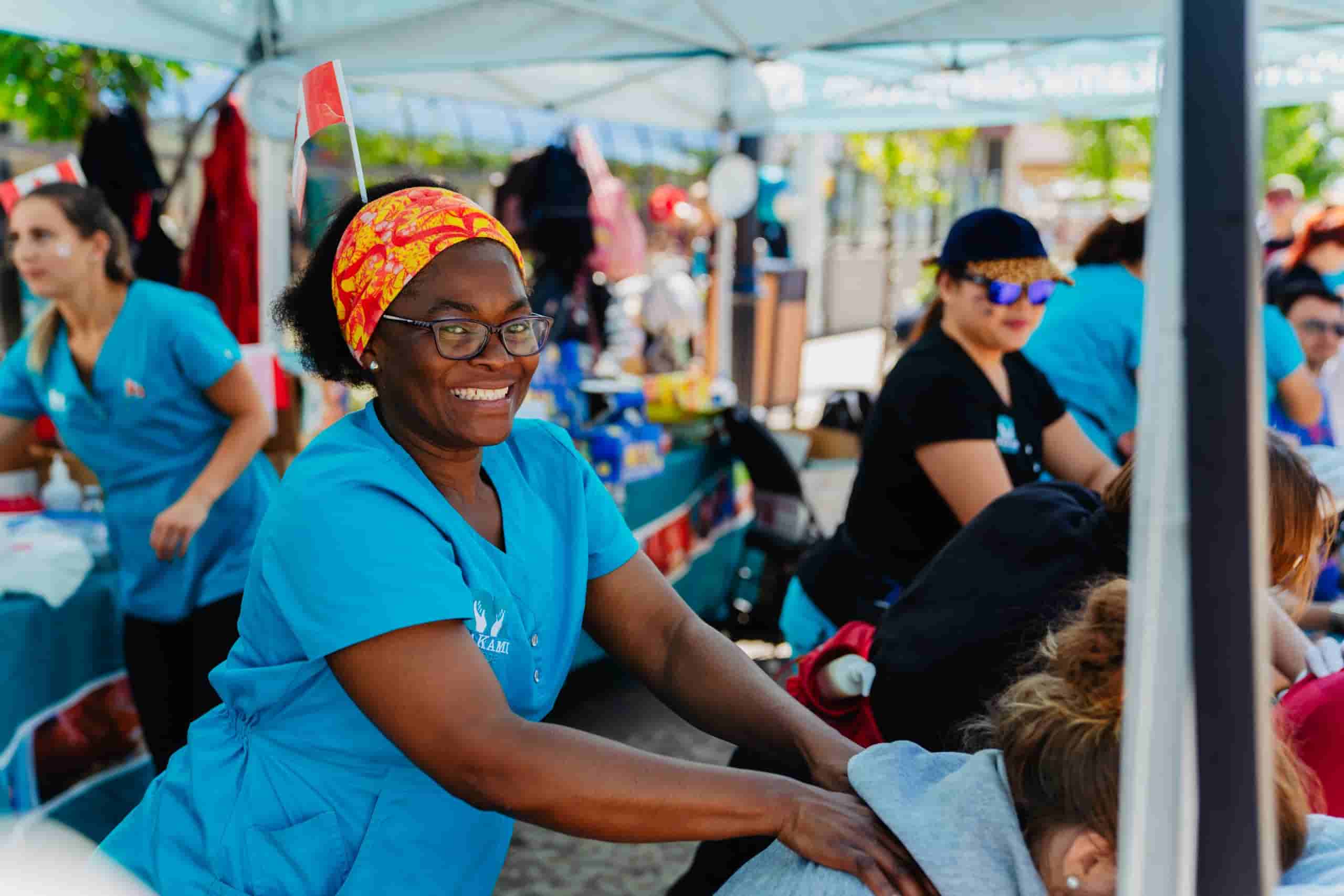 A middle aged black female student wearing a teal blue scrub top with the MaKami College logo on it massages someone in a seated chair while volunteering at an outreach event for the post-secondary college.