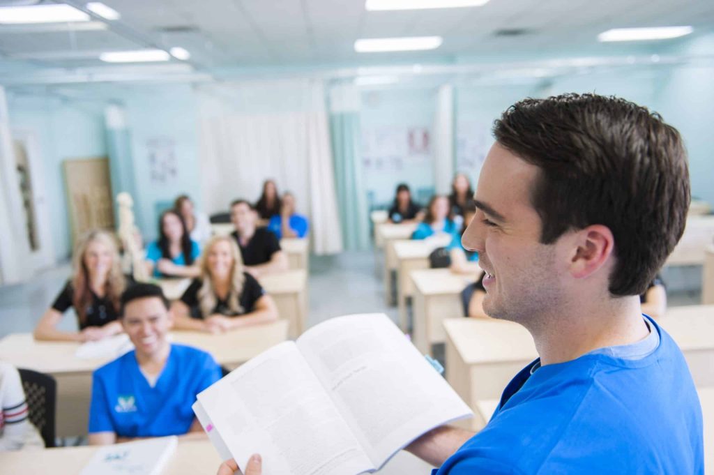 A young male student is featured in the foreground holding a book while lecturing to students in the background of MaKami College