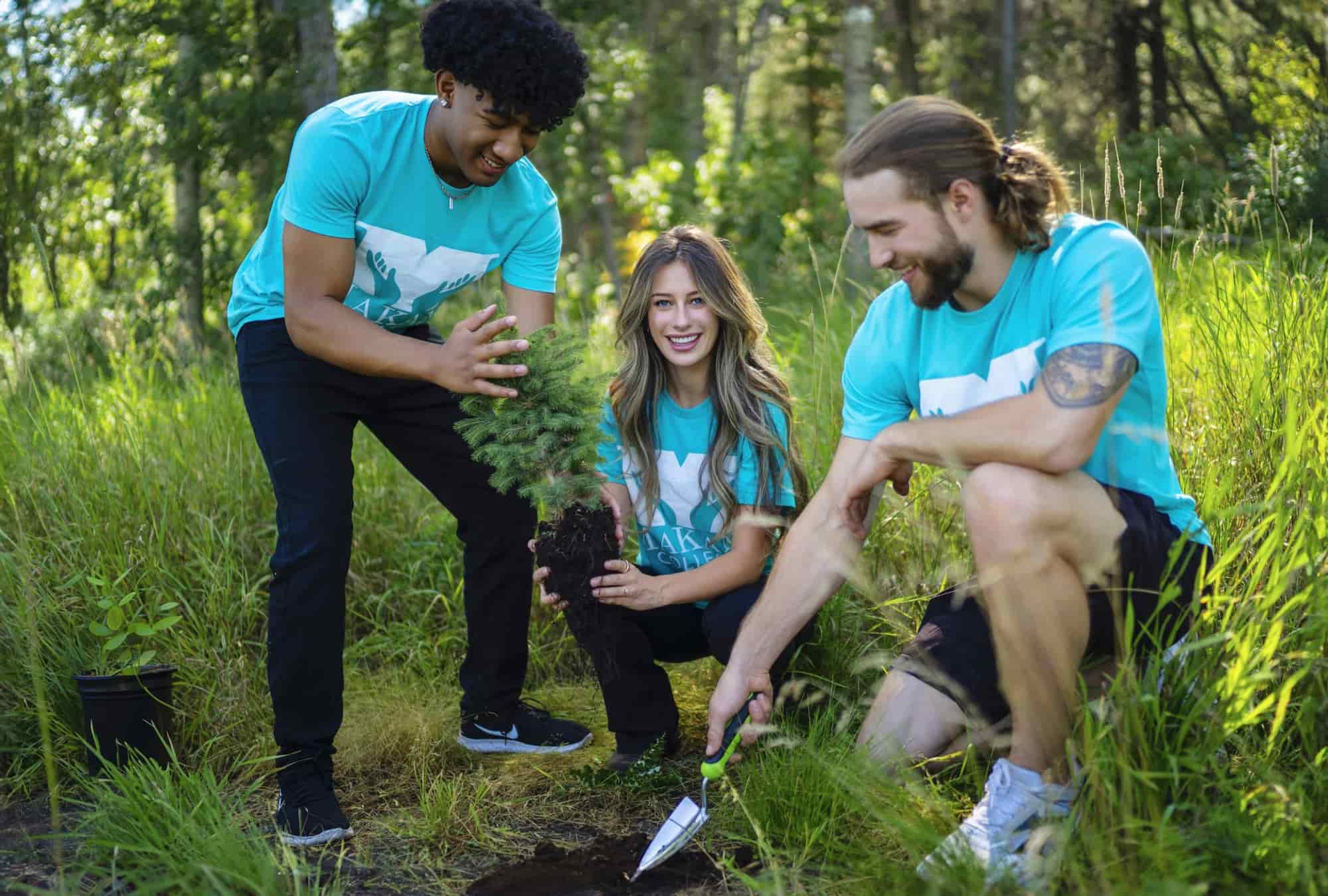 A young African male on the left holds a spruce tree to plant, next to a young caucasian woman and young caucasian male in Red Deer, Alberta, Canada