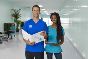 A tall, white caucasian male stands holds a white MaKami binder standing next to an African Canadian woman in the brightly lit hallways of MaKami College