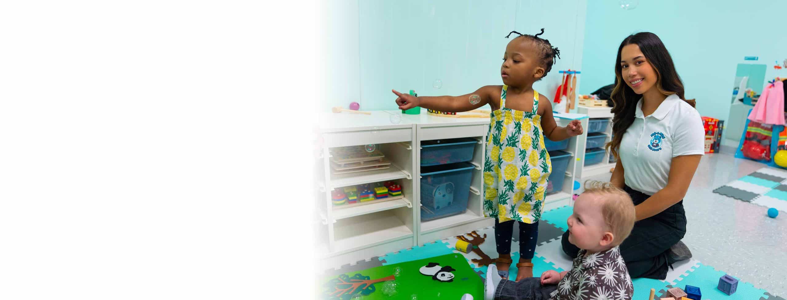 A young dark haired ethnic early childhood educator smiles at the camera as a young african canadian female toddler points to something on the floor inside an early childhood education setting