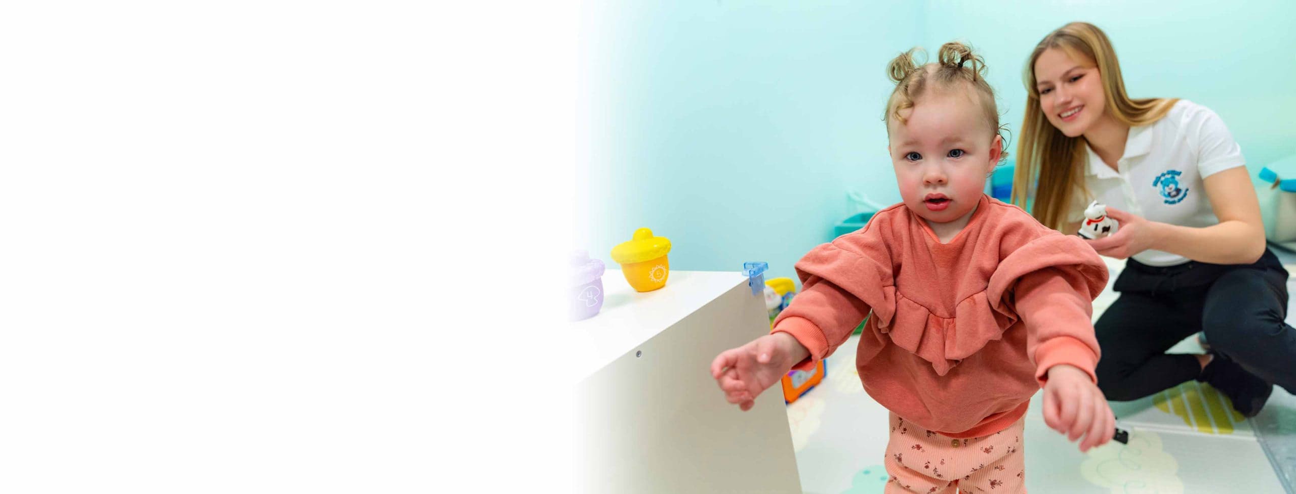 A young female caucasian child walks with her hands outstretched to the camera while a young female childcare worker smiles behind her