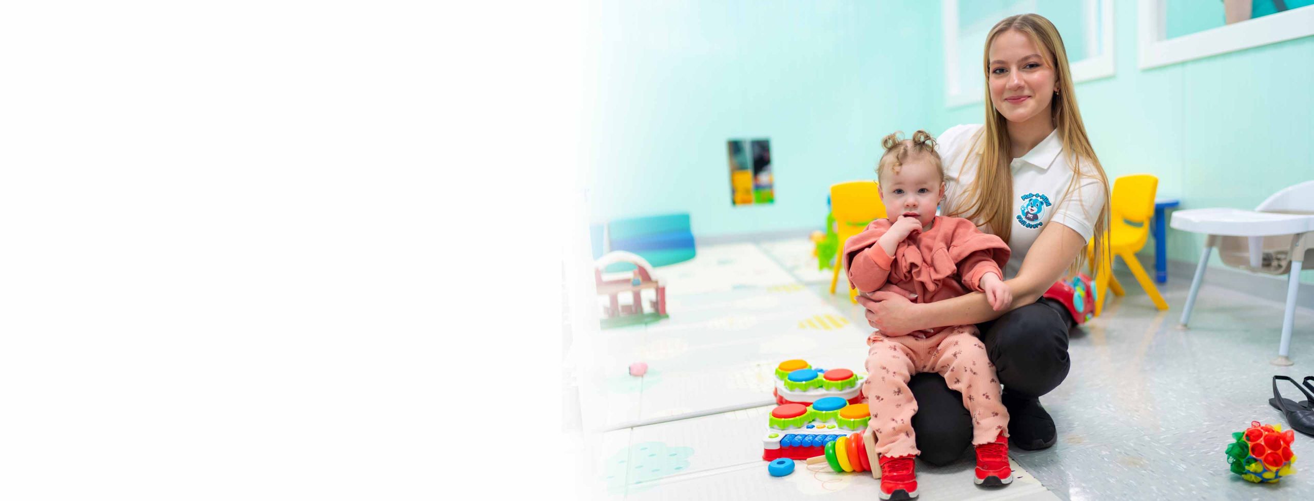A young blonde caucasian female early childhood educator sits with a young caucasian female toddler in an early childhood education setting
