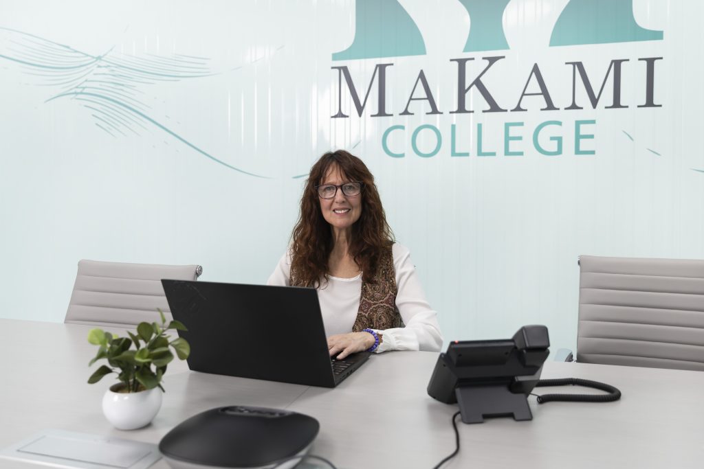 Allison Taylor, a middle aged caucasian woman with long dark wavy hair and glasses sits behind her laptio wiht a phone next to her and the MaKami College logo behind her at MaKami College in Calgary, Alberta, Canada.