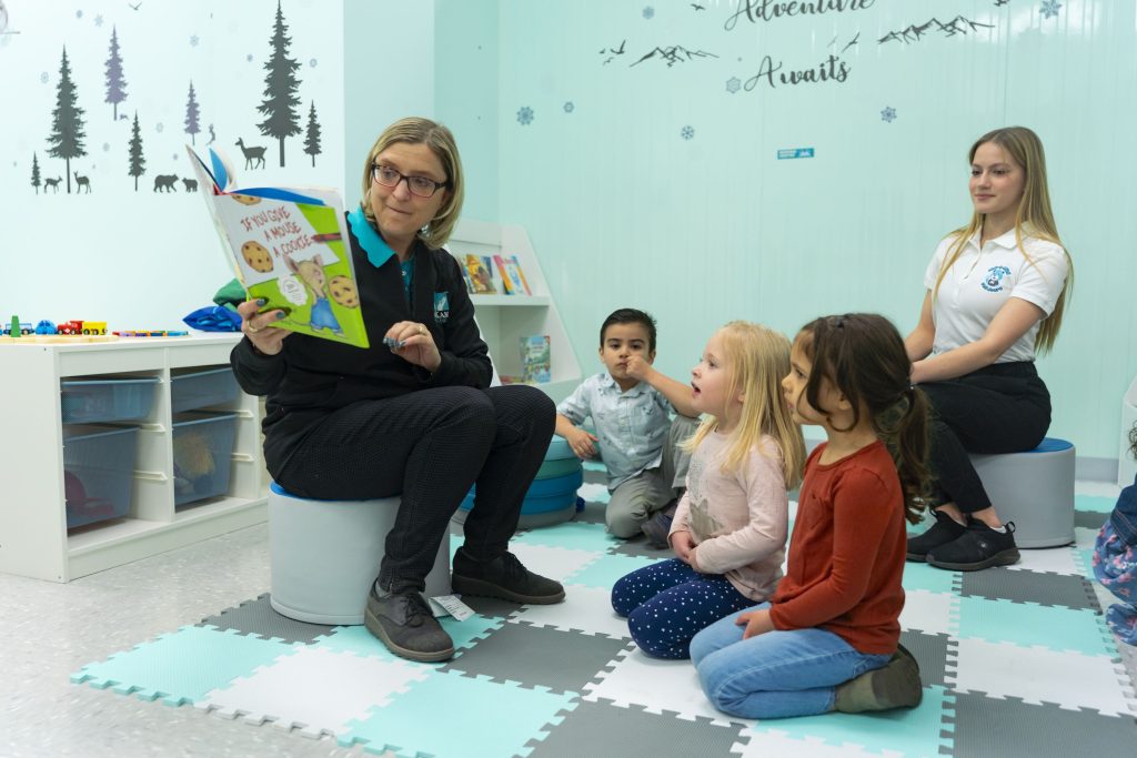 A caucasian middle aged female with short blong hair and glasses holds a book up in front of young children in an early childcare setting