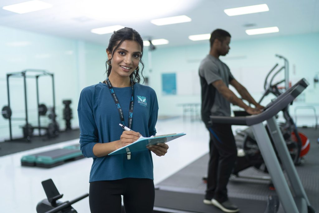 A young dark haired Indian woman looks at the camera while holding a clipboardin a gym, while a young African man walks on a treadmill in the background