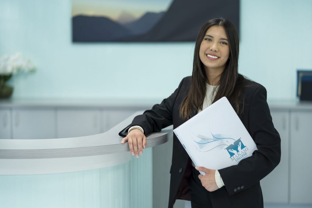 Smiling business student holding MaKami College folder at a modern reception desk, promoting MaKami College's Business Administration program in Alberta