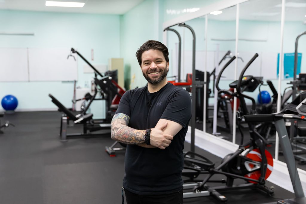 Personal trainer smiling with crossed arms in MaKami College's fitness center with stationary bikes and strength training equipment