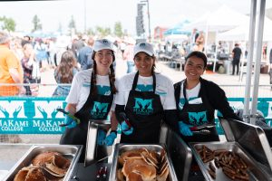 Three people in white shirts, black 'Makami College' aprons, and blue gloves serve food from trays at an outdoor event, with a crowd and trees in the background.