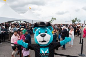 A blue bear mascot in a cowboy hat and 'MAKAMI COLLEGE' shirt stands with arms outstretched at a large outdoor event under white tents, surrounded by attendees.