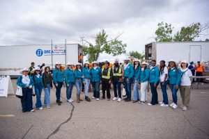 Group of people in blue shirts and white hard hats, some in safety vests, standing outdoors near two large trucks labeled 'DREAM,' with trees and a cloudy sky in the background.