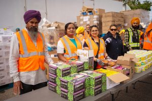Group of people in orange safety vests stand behind a table with boxes and containers of food, with stacks of supplies in the background.