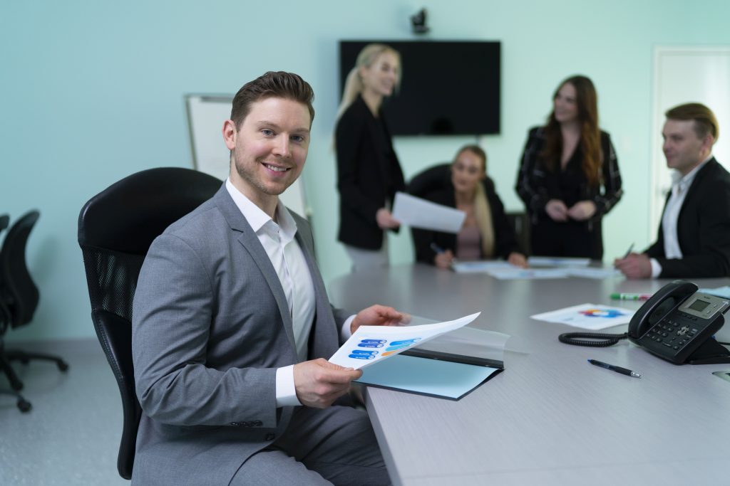 A group of five people in business attire are gathered in a conference room. One person is seated at the head of the table holding a document with charts, while the others are standing or sitting around the table, also holding papers. The room has a large screen on the wall and a conference phone on the table.
