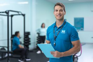 A person wearing a blue polo shirt and an ID lanyard around the neck standing in a gym holding a blue clipboard. In the background, there are two other people, one sitting on an exercise bench and another standing near weight racks. The gym has light blue walls and various fitness equipment.