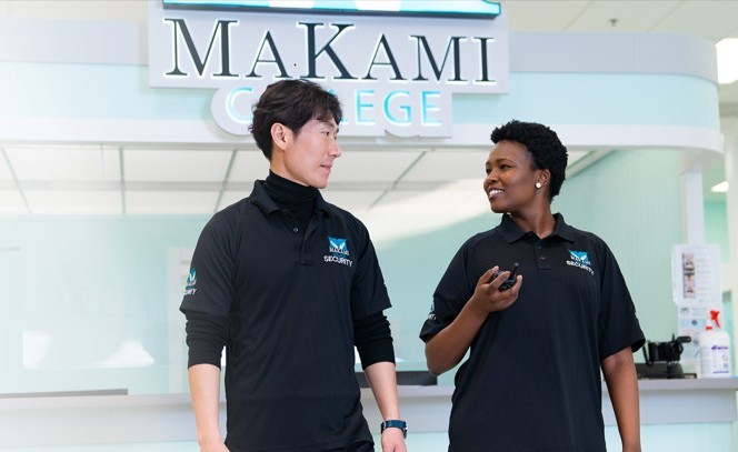 Two security personnel in black uniforms labeled 'SECURITY' stand in front of a reception desk at MaKami College. The background features a light blue and white interior with the college's name displayed above them.