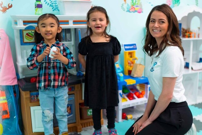 Two children and an adult are in a playroom. One child wears a plaid shirt and jeans, holding a toy. The other wears a black dress. The adult, kneeling beside them, wears a white shirt and black pants. Toys and play kitchen equipment are visible in the background.