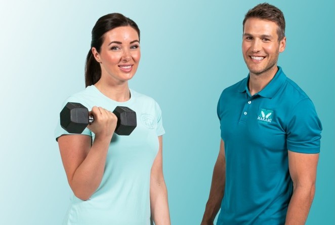 Two individuals standing side by side against a light blue background. One is holding a black dumbbell and wearing a light blue shirt; the other is in a teal polo shirt.