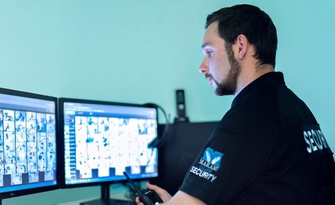 A security guard monitors multiple surveillance camera feeds on two computer monitors. The guard wears a black uniform labeled 'SECURITY' with a 'MAKAMI SECURITY' logo and holds a walkie-talkie.