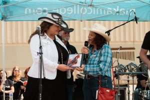 Under a blue tent labeled 'makamicollege.com,' a person in a white jacket and black dress receives an award from another individual in a blue plaid shirt and jeans. Musical instruments and other people are visible in the background.