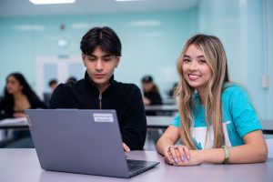 Two students are sitting at a desk in a classroom, working together on a laptop. The classroom has several other students in the background, also engaged in their work. The room is well-lit with light blue walls and large windows.