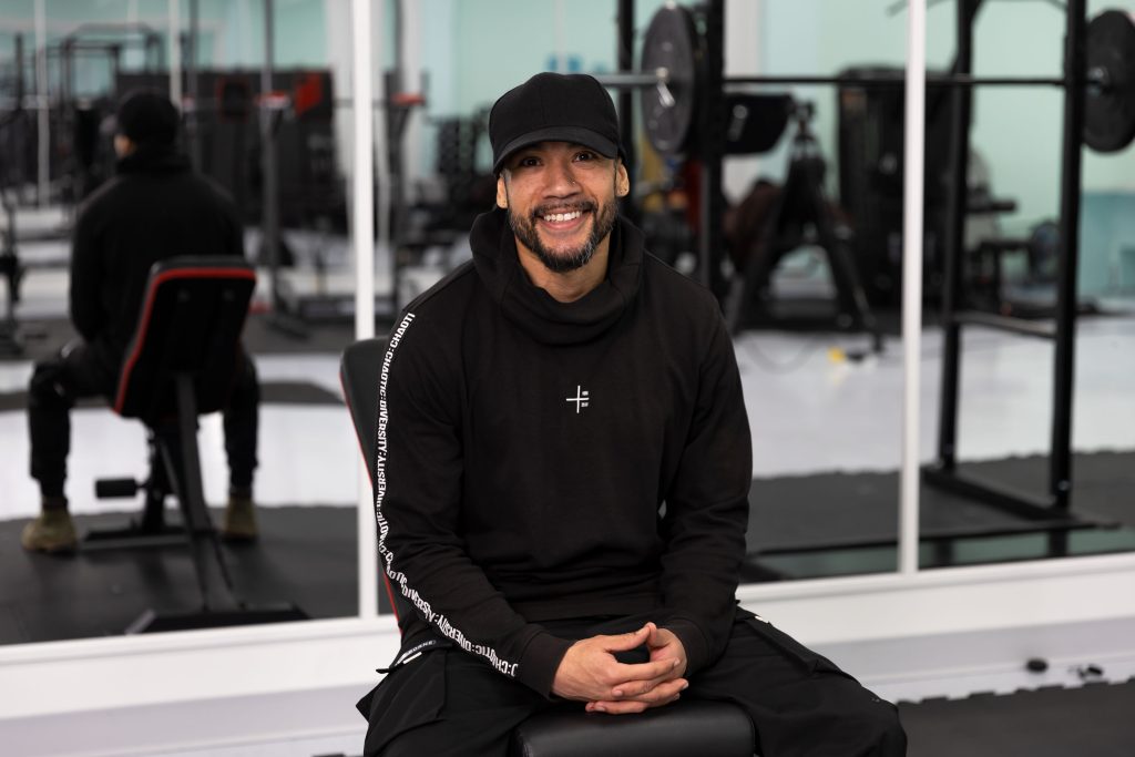 Person sitting on a bench in a gym, wearing a black hoodie with white text. Gym equipment and mirrors in the background.
