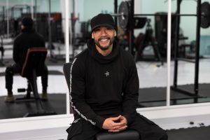 Person sitting on a bench in a gym, wearing a black hoodie with white text. Gym equipment and mirrors in the background.