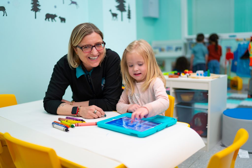 Child using a tablet at a classroom table with colorful markers, while an adult provides guidance in an early learning environment.