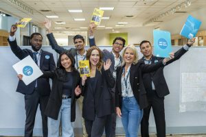 Group of people standing indoors in a bright office space, holding up colorful pamphlets and folders with messages like ‘Get Out and Vote’ and ‘Ready, Set, Vote.’ They are dressed in business attire and appear to be celebrating or promoting a voting campaign.