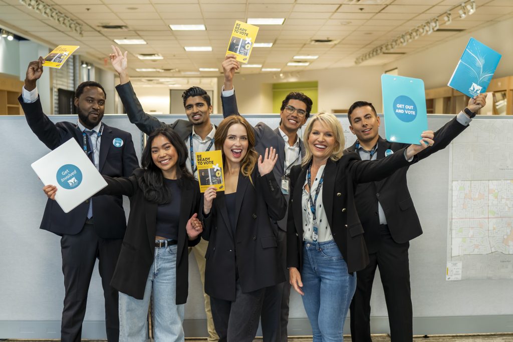 Group of people standing together in an office setting, holding up informational pamphlets and folders with messages such as ‘Get Out and Vote.’ They are dressed in business-casual attire, wearing lanyards, and appear to be promoting civic engagement and voter participation.