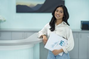 Person standing at a modern reception desk holding a white folder with a blue logo and abstract design, in a bright office setting with light-colored walls and cabinets in the background.