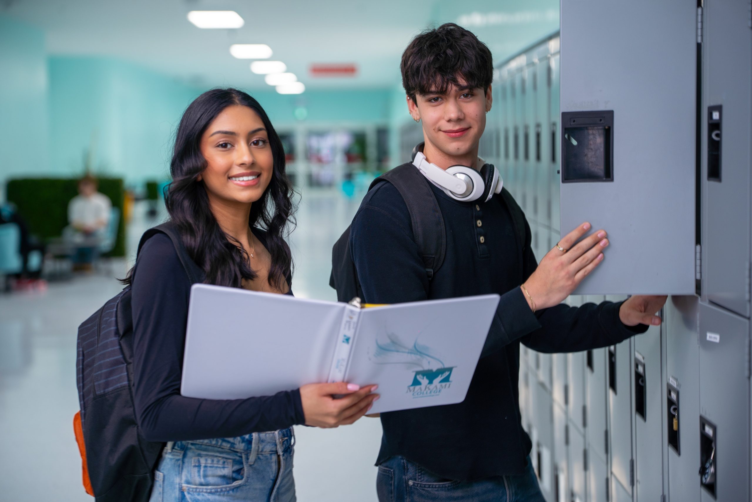 Two students standing by lockers in a school hallway, one holding an open binder and the other opening a locker.