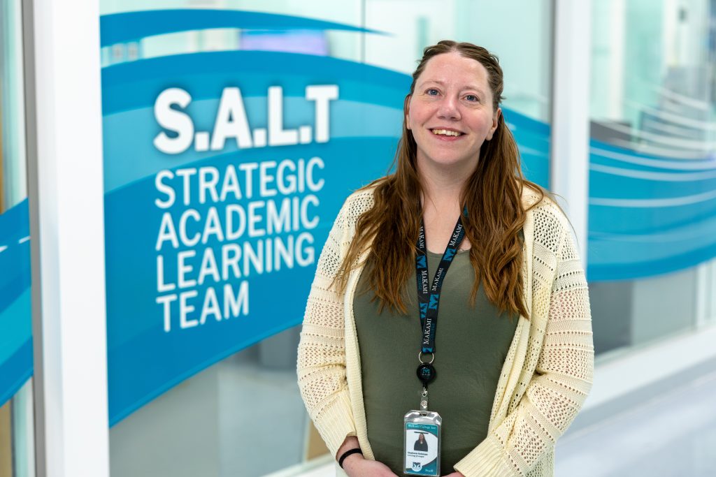 A person with long wavy hair stands in front of a blue wall graphic that reads “S.A.L.T Strategic Academic Learning Team.” They are wearing a light cream knit cardigan over a green top and have a MaKami-branded lanyard with an ID badge around their neck.