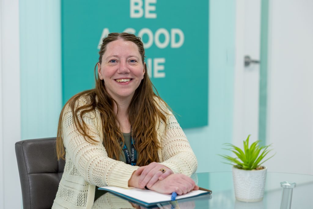 A staff member seated at a glass table in a modern MaKami College office, wearing a light knit cardigan and lanyard, with a notebook and pen in front of them and a small potted plant on the table. A teal wall sign in the background reads "Be a Good One."