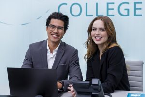 Two professionals sitting at a modern office desk with a laptop and phone, collaborating in a college reception or administrative setting.