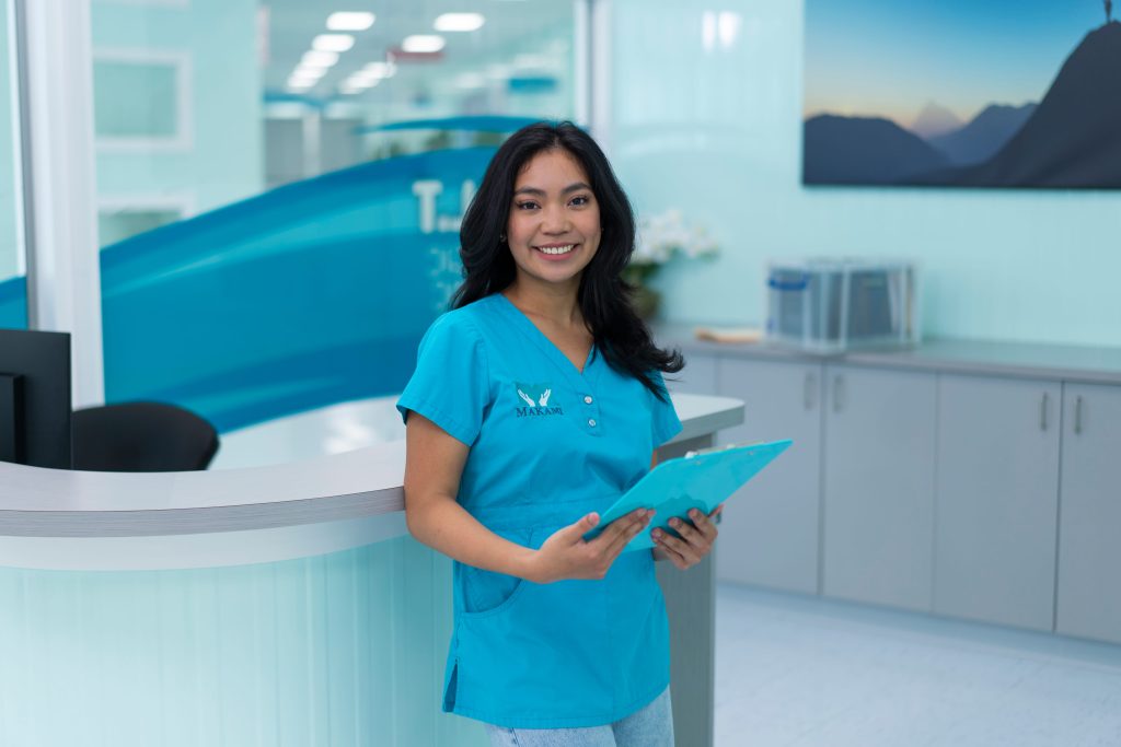 Smiling MaKami College medical office assistant student in scrubs assisting a doctor with a patient chart in a clinical setting
