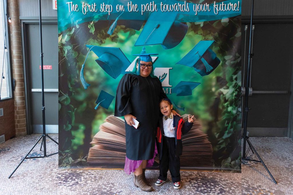 Graduate in black cap and gown holding diploma poses with young child in front of MaKami College graduation backdrop reading "The first step on the path towards your future!" at Shania Stevens' MaKami College graduation ceremony