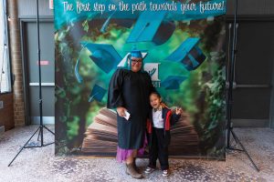 Graduate in black cap and gown holding diploma poses with young child in front of MaKami College graduation backdrop reading "The first step on the path towards your future!" at Shania Stevens' MaKami College graduation ceremony